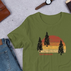 Puede incluir: Una camiseta verde con un gráfico de puesta de sol de estilo vintage que presenta tres árboles en silueta y el texto "Get Outside".