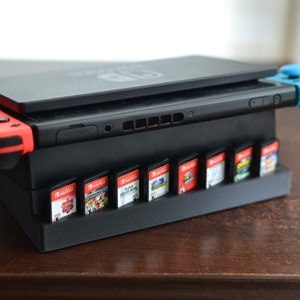 May include: A black Nintendo Switch console with a red and blue Joy-Con controller on each side. The console is sitting on a black stand with a built-in game cartridge holder. The stand is holding eight game cartridges.