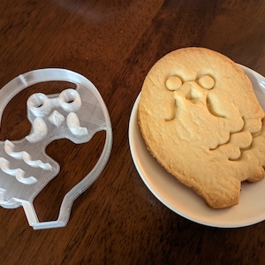 Peut inclure: Un emporte-pièce en plastique blanc en forme de fantôme avec un visage et un biscuit blanc cuit en forme d'emporte-pièce.