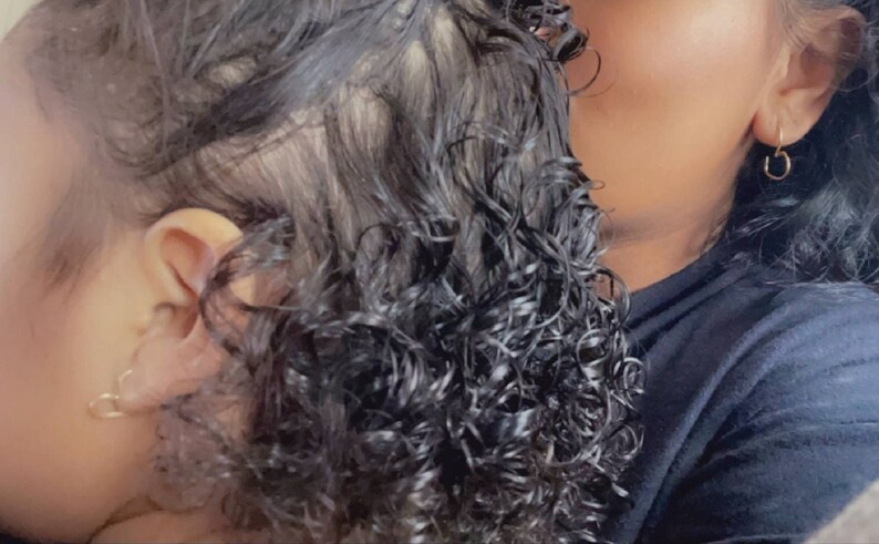 May include: A close-up of a young child with curly black hair, wearing a small hoop earring in their left ear. The child is being held by an adult with dark hair, who is wearing a gold hoop earring in their right ear.