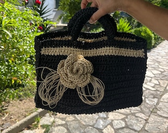 Könnte beinhalten: Eine schwarze gewebte Handtasche mit einer beigen Blume und Zierleiste. Die Tasche hat zwei Griffe und eine rechteckige Form. Die Blume besteht aus gewebtem Material und hat lange, dünne Strähnen, die sich davon erstrecken. Die Tasche wird von einer Person gehalten.