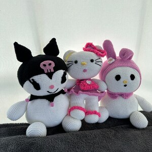 Puede incluir: Tres juguetes de peluche de crochet: un gato blanco y negro con una calavera en el pecho, un gato blanco con orejas rosas y un vestido rosa, y un conejito rosa con un lazo rosa. Los juguetes están sentados sobre una superficie gris.