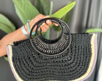 Könnte beinhalten: Eine schwarze gehäkelte Tragetasche mit helllila und beigefarbenem Besatz. Die Tasche hat zwei große, runde, schwarze geflochtene Henkel.