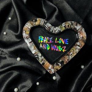 May include: Heart-shaped trinket tray with a black interior and a border of gold and silver flakes. The tray features the text "PEACE LOVE AND WUBZ" in a rainbow holographic font. The tray sits on a black satin surface.