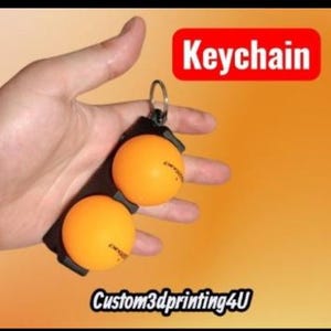 May include: A keychain featuring two orange ping pong balls. The balls are attached to a black strap with a metal ring. The word "Keychain" is in a red box. The text "Custom3dprinting4U" is at the bottom.