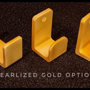 May include: Three gold-colored wall hooks with a curved design. The hooks are made of metal and have a pearlized finish.  PEARLIZED GOLD OPTION is written in black text at the bottom of the image.