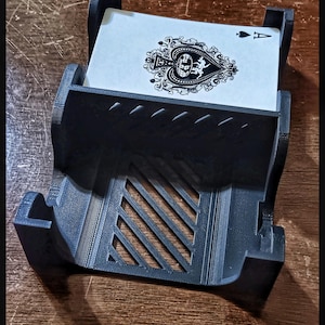May include: A black plastic card holder with a decorative design. The holder is designed to hold a single playing card. The card in the holder is the Ace of Spades.