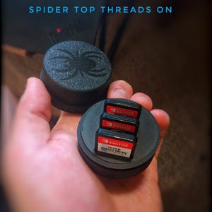 May include: A black plastic case with a spider design on the top. The case holds four Nintendo Switch game cartridges. The cartridges are labeled "Nintendo Switch" and "Super Smash Bros. Ultimate".