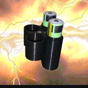 May include: Black 3D-printed battery holder with two cylindrical slots for batteries and two additional slots. Two green and silver batteries are inserted. The holder is designed to organize and store batteries.