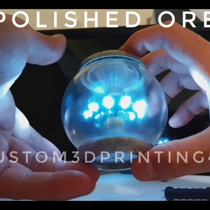 Könnte beinhalten: Eine klare Glaskugel mit einem blauen Licht im Inneren, gehalten in zwei Händen. Die Kugel sitzt auf einem Sockel mit silbernem Finish. Der Text "CUSTOM3DPRINTING4U" ist am unteren Rand des Bildes sichtbar.