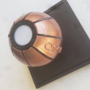 May include: A copper-colored sphere with a circular opening and a raised inscription that reads "OSG". The sphere is on a dark brown base.