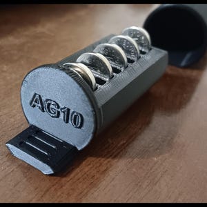 May include: A black 3D-printed battery holder with the text "AG10" on the end. The holder contains several silver AG10 button cell batteries. The cylindrical cap is open, revealing the batteries inside the rectangular compartment.