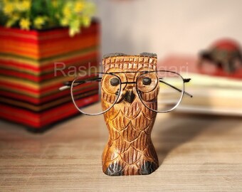 May include: Wooden owl figurine with a carved design, wearing a pair of black framed glasses. The owl is brown and has a detailed beak and eyes.