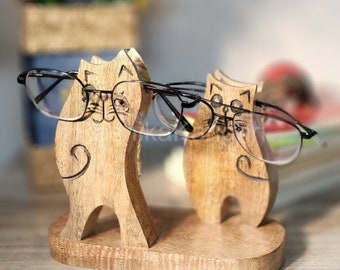 May include: Wooden cat figurine eyeglasses holder with two cats wearing glasses. The cats are brown and have black eyes. The glasses are black and have a silver frame.