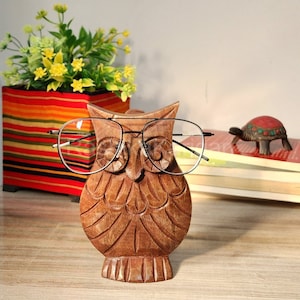 May include: Wooden owl figurine with black glasses, used as a glasses holder. The owl is brown and has a detailed carving. It is sitting on a wooden surface.