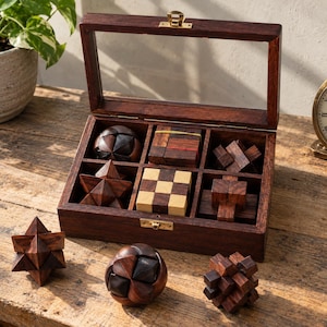 May include: A dark brown wooden storage box with a hinged glass lid containing six distinct wooden brain teaser puzzles. Three additional wooden puzzles are arranged in front of the box, featuring various geometric shapes including spheres and stars.