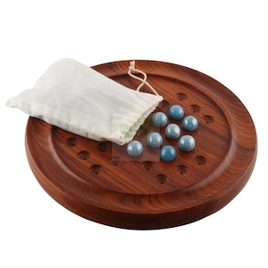 May include: A wooden solitaire game with a dark brown, circular board. The board has evenly spaced holes and a set of blue marbles. A white drawstring bag rests on the board, next to the marbles.