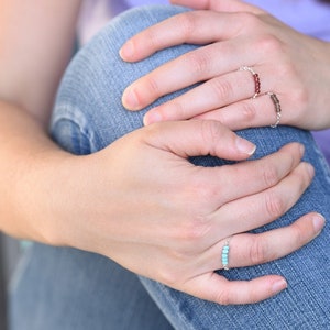 May include: Three beaded rings on fingers. One ring features small, light blue beads, another has dark red beads, and the third has brown beads. The rings are on a silver chain. The person is wearing blue jeans.