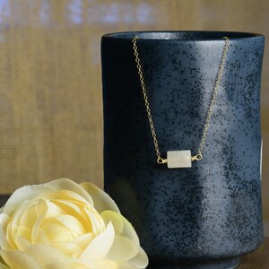 May include: A delicate gold chain necklace with a white rectangular stone pendant. The necklace is displayed on a dark blue ceramic mug and a white flower.