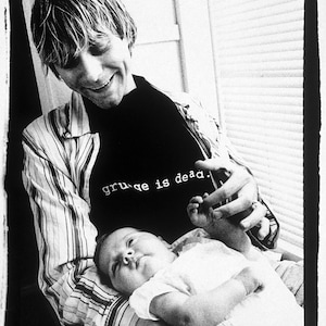 May include: A black and white photo of a man holding a baby. The man is wearing a striped shirt and a black t-shirt with the text "grunge is dead." The baby is wearing a white onesie.