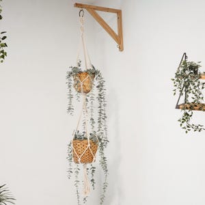 Peut inclure: Deux jardinières suspendues en macramé, accrochées à un support en bois. Des plantes vertes retombantes s'échappent des paniers, créant une décoration naturelle. L'ensemble est posé sur un mur blanc.