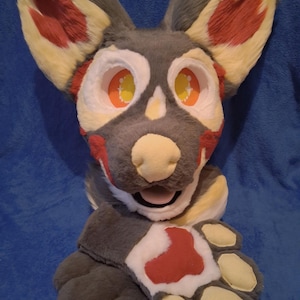 May include: A plush, anthropomorphic animal head and paw set. The head features large, expressive eyes with orange and yellow details, gray fur, and red accents. The paw has gray fur with a white and red paw pad.