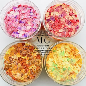 May include: Four jars of iridescent glitter in various shades of pink, orange, and yellow. The glitter is shaped like seashells. The jars are labeled "Bright Pink Pearl", "Coral Pink", "Golden Amber", and "Bright Sunny Day".