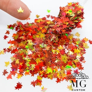 May include: A close-up of iridescent holographic glitter shaped like maple leaves in shades of red, orange, yellow, and green. The glitter is scattered on a white surface and a small scoop of glitter is being held by a finger.