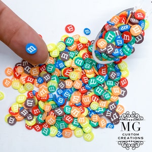 May include: A large assortment of colorful round candy sprinkles with the letter "M" printed in white on each sprinkle. The sprinkles are scattered on a white surface and in a spoon.