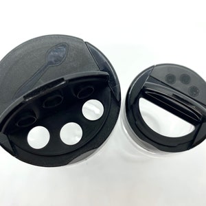 May include: Two black plastic spice jar lids with a hinged opening and a small spoon inside one lid. The lids have multiple holes for dispensing spices.