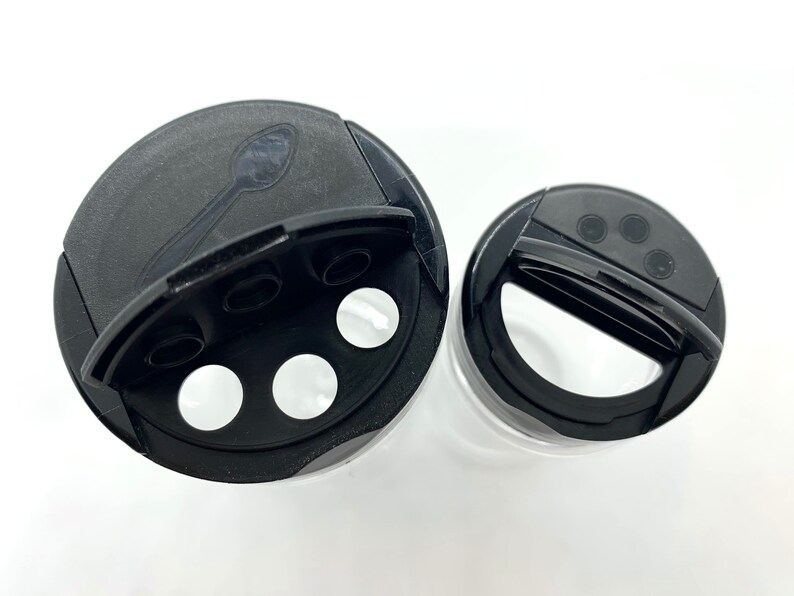 May include: Two black plastic spice jar lids with a hinged opening and a small spoon inside one lid. The lids have multiple holes for dispensing spices.