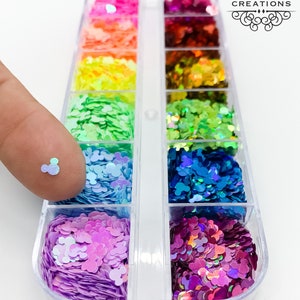 May include: A clear plastic container with 12 compartments filled with iridescent glitter in various colors. Each compartment contains a different color of glitter, including pink, purple, blue, green, yellow, orange, and red. The glitter is shaped like Mickey Mouse ears.