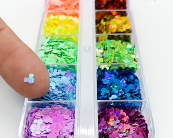 Puede incluir: Un recipiente de plástico transparente con 12 compartimentos llenos de purpurina iridiscente en varios colores. Cada compartimento contiene un color diferente de purpurina, incluyendo rosa, morado, azul, verde, amarillo, naranja y rojo. La purpurina tiene forma de orejas de Mickey Mouse.