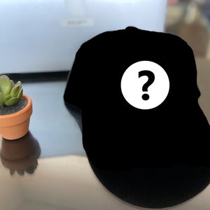 Może przedstawiać: Czarna czapka z daszkiem z białym znakiem zapytania w kółku z przodu.