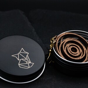 May include: Wooden spiral earrings with gold-colored hooks rest in a silver tin. The tin is next to a black container with a geometric fox logo. The earrings have a layered, circular design and are made of a dark wood.