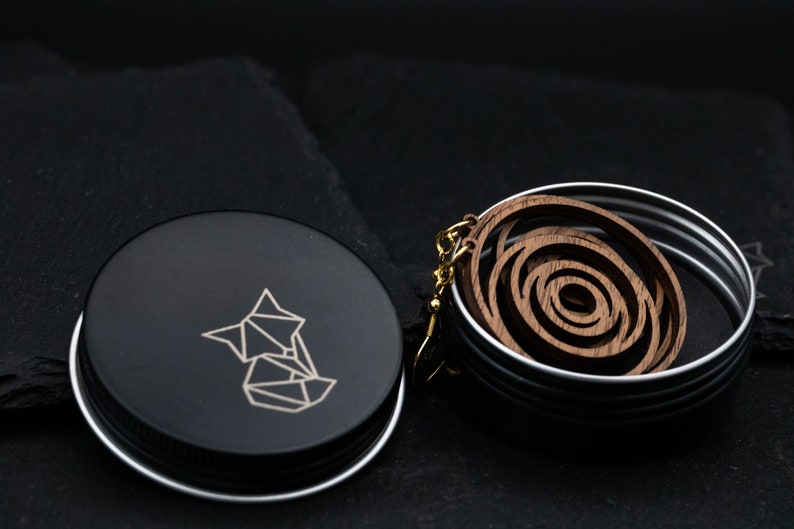 May include: Wooden spiral earrings with gold-colored hooks rest in a silver tin. The tin is next to a black container with a geometric fox logo. The earrings have a layered, circular design and are made of a dark wood.