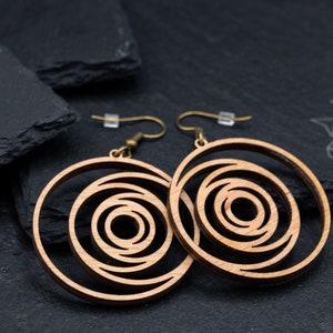 May include: Two wooden spiral earrings with brass-colored hooks. The earrings have a light brown color and a layered, concentric design. The background is a dark gray surface.