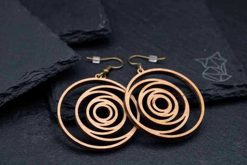 May include: Two wooden spiral earrings with brass-colored hooks. The earrings have a light brown color and a layered, concentric design. The background is a dark gray surface.