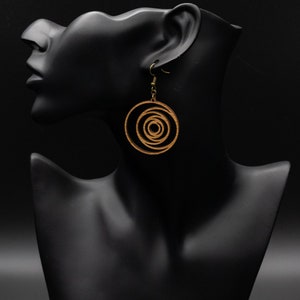 May include: Wooden spiral earrings with brass-colored hooks hang from a black mannequin head. The earrings feature a concentric circle design, with a light brown color. The background is solid black.