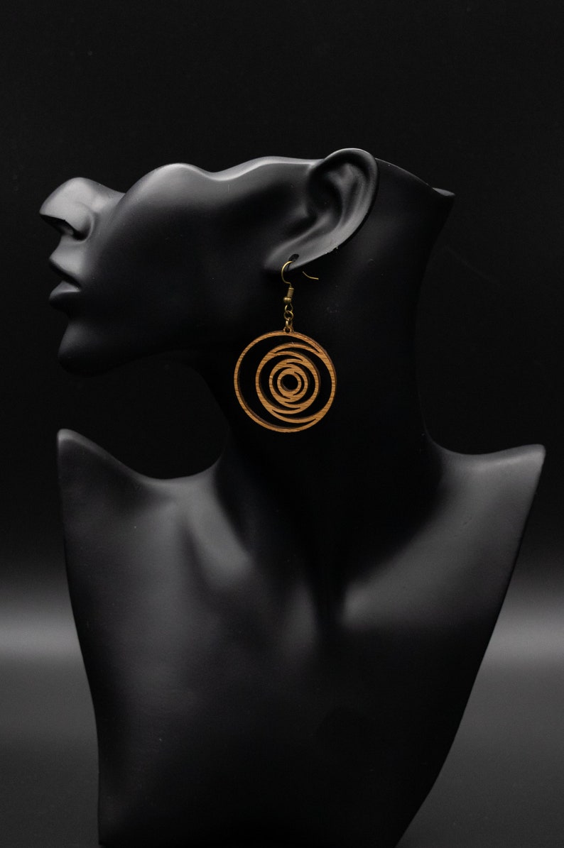 May include: Wooden spiral earrings with brass-colored hooks hang from a black mannequin head. The earrings feature a concentric circle design, with a light brown color. The background is solid black.