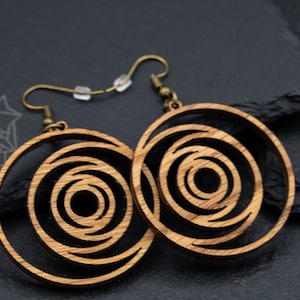 May include: A pair of wooden earrings with a spiral design. The earrings are circular with concentric circles cut out, creating a layered effect. They have bronze-colored ear wires and are displayed on a dark gray surface.