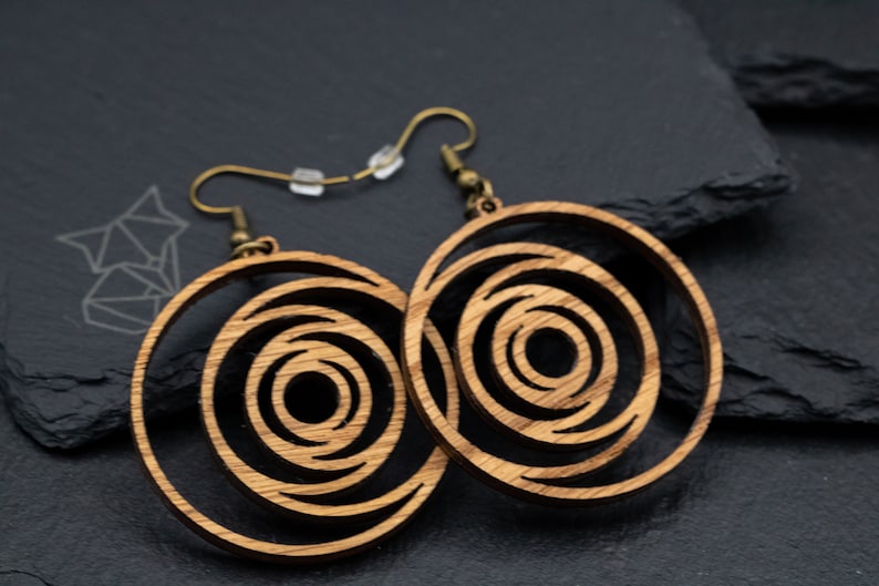 May include: A pair of wooden earrings with a spiral design. The earrings are circular with concentric circles cut out, creating a layered effect. They have bronze-colored ear wires and are displayed on a dark gray surface.