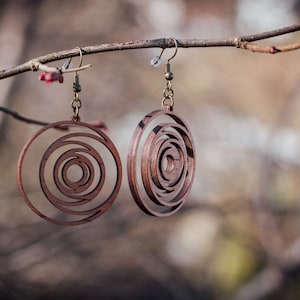 May include: Two wooden earrings with a spiral design hang from a tree branch. The earrings are a warm brown color and have a circular shape with concentric circles cut out. The earrings are suspended by bronze-colored hooks.