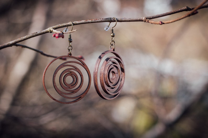May include: Two wooden earrings with a spiral design hang from a tree branch. The earrings are a warm brown color and have a circular shape with concentric circles cut out. The earrings are suspended by bronze-colored hooks.