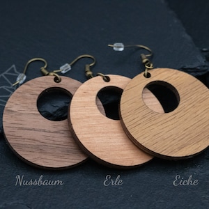May include: Three wooden hoop earrings in varying shades of brown, with a circular cutout in the center. The earrings are suspended from bronze-colored ear wires. The earrings are labeled "Nussbaum", "Erle", and "Eiche".
