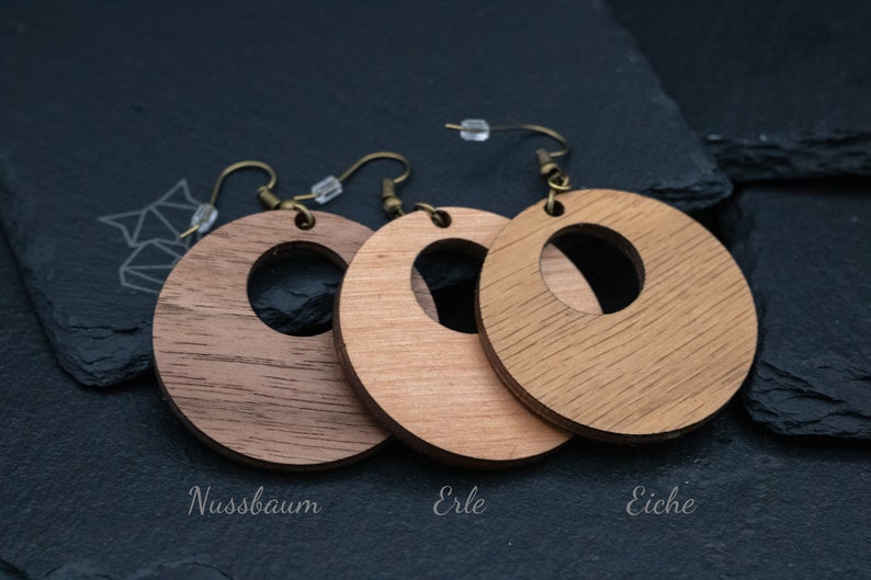 May include: Three wooden hoop earrings in varying shades of brown, with a circular cutout in the center. The earrings are suspended from bronze-colored ear wires. The earrings are labeled "Nussbaum", "Erle", and "Eiche".