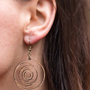 May include: Wooden earrings with a concentric circle design. The earrings are a light brown color and hang from a small, gold-colored hook. The design features multiple circular layers, creating a spiral effect.