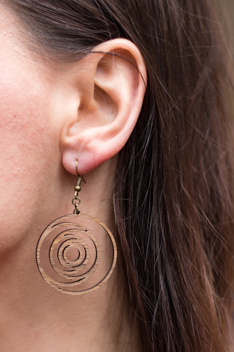 May include: Wooden earrings with a concentric circle design. The earrings are a light brown color and hang from a small, gold-colored hook. The design features multiple circular layers, creating a spiral effect.