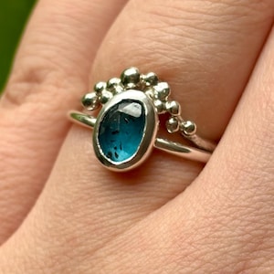 Può includere: Un anello d'argento con una gemma ovale color teal e un gruppo di piccole perle d'argento. L'anello è su una mano con uno sfondo verde sfocato.