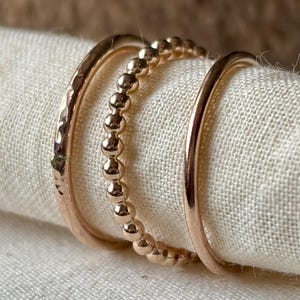 May include: Three gold rings on a white cloth. The rings feature distinct designs: a hammered texture, small beads, and a smooth, rounded band. The rings are probably made of gold or a gold-coloured metal. The cloth is a neutral colour.